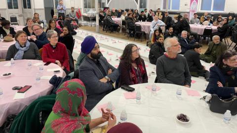people from different faiths sit at tables in a large hall looking towards a stage 
