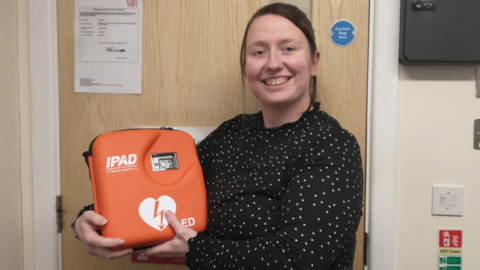 Naomi, wearing a black blouse with small white spots, smiling to camera, while holding the orange defibrillator pack in front of a fire door