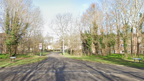 A wide tree-lined road with a sign at the entrance saying "Deerleap" with blocks of flat in the background. 