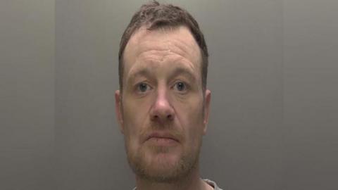 Police custody photograph of Timothy Tinson. He has short brown hair and beard and is wearing a grey T-shirt