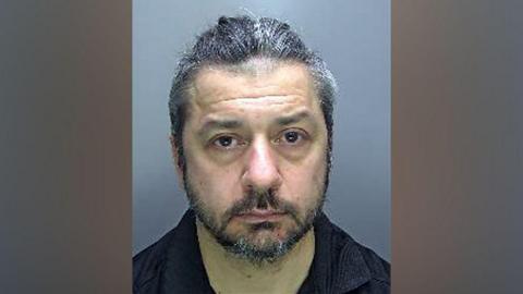 Man with greying hair and facial hair in a police custody photo.
