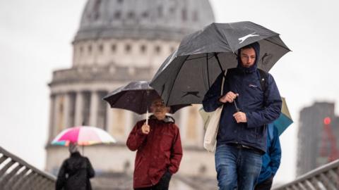 People walking across a bridge holding umbrellas and wearing raincoats with the blurred image of St Paul's Cathedral in the background