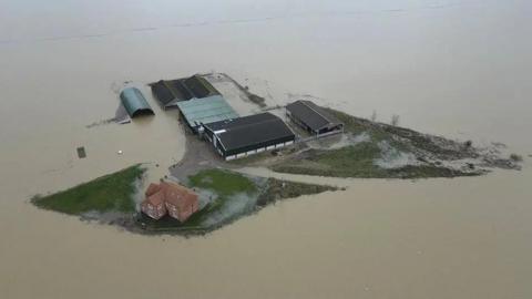 A farmhouse on slightly elevated ground is completely surrounded by muddy flood water.