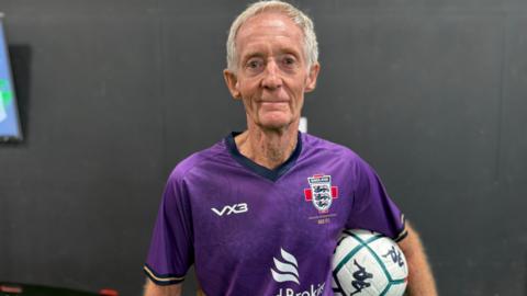 Geoff Martel is wearing a purple shirt with the England logo which features three blue lions. He is holding a white football with green lines on it under his left arm. he has short white hair and is looking straight at the camera.