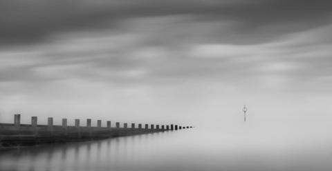 A black‑and‑white scene showing a long wooden pier stretching diagonally into calm, misty water. The sky is cloudy and blurred, and a single navigation marker stands alone in the distance.