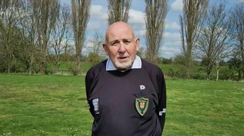 Ernie Broughton, wearing a black referee's kit, looks into the camera lens. His hands are behind his back and he has a stern look. 