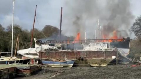 A picture of the fire which has taken over the boat in the estuary. There is large orange flames.