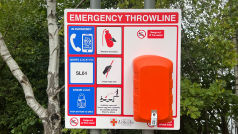 A wide-shot image of an emergency throwline station. It is bright blue, orange and red. There are green trees behind.