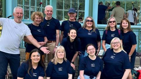 Ten people in navy blue T shirt and one man in a white T shirt pose in front of a teal bandstand. They are smiling at the camera.