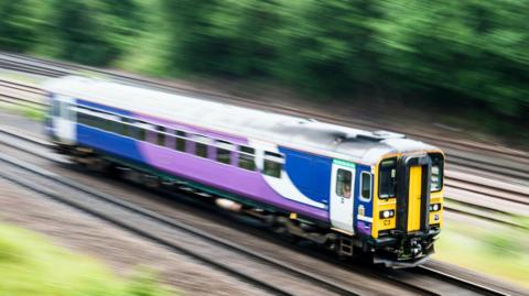A blue, white and purple train, part of the Northern Trains service, pictured mid-journey.
