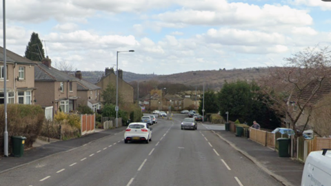 A suburban street lined with houses, cars, and hills in the background