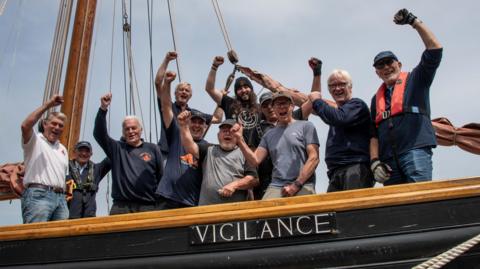 A crew of 11 men raise their hands in the air on board a boat called Vigilance.
