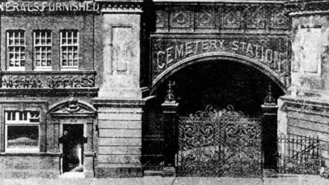 A black and white image of a building with an ornate gate. The gate has the words "Cemetery Station" above it.