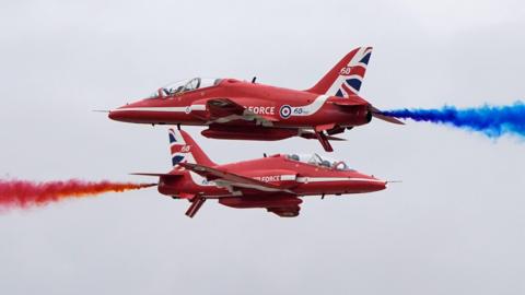 The Red Arrows display team crisscrossing, with red and blue smoke billowing from behind them.