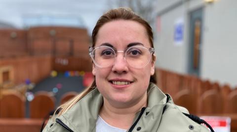 India Chapman Webb is smiling at the camera as she has her brown hair tied back in a ponytail and is wearing glasses. She is also wearing a green raincoat and a white top.