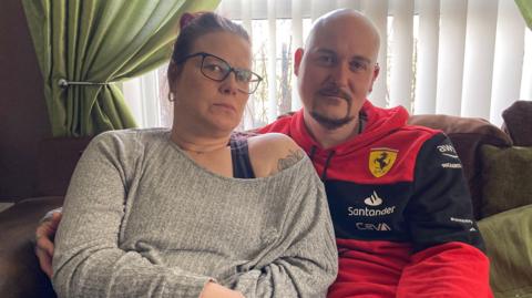 A man and woman sitting on a brown sofa, in a room with green curtains. They both have serious expressions. She is wearing a grey long-sleeved top revealing a floral tattoo on one shoulder and has brown hair tied back and brown glasses. He is wearing a red hoodie with a Ferrari logo on it, has a shaved head and brown beard. 