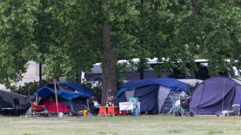 A general view of a homeless camp in Park lane. There are five tents in shot and two people with blurred faces are seated doing what appears to be eating around a table. A white coach with blacked windows is pictured in the background and in-between the tents is a tree trunk. 