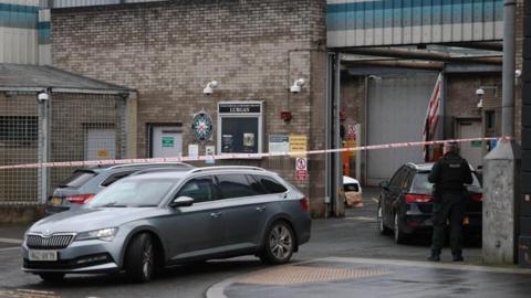 Two grey unmarked police cars sit in front of a highly fortified police station. There is a also a black unmarked police car, a police officer standing with his back to camera and a cordon in place.