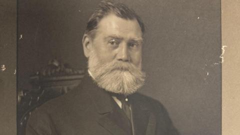 A black and white photo of Frederick Sutton, sitting on a carved wooden chair. He has a large white beard, short hair and wears a smart suit of the period.