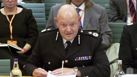 Craig Guildford at a select committee meeting. He is sitting in uniform at a desk, with small white microphone in front of him. In the background are a handful of people on green seats