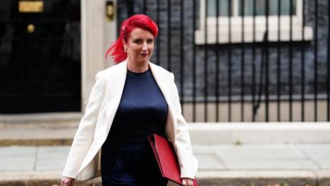 Woman with red hair wearing a blue dress and white jacket holds a red floder in her left hand as she walks out of the black door at Number 10 Downing Street.