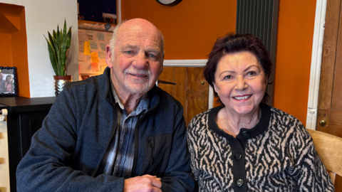 A man with thin grey hair and a grey beard with a blue jacket and blue checked shirt sitting beside a woman with dark hair and a black and white patterned top in a room with orange walls