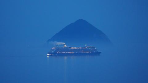 A cruise ship, with lots of lights and a large plum of smoke coming from the top sailing in front of Ailsa Craig, a large dark rock in the water. The ship and rock are the only thing visible as a dark blue sky and water merge.