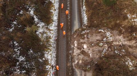 An aerial view of a railway track with a pile of earth over the track on the right, following a landslip. Workers wearing bright orange outfits can be seen working on the track.