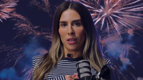 BBC presenter looks at camera. She is wearing a black and white stripe top and is speaking in to a microphone. There is an image of fireworks in the background.