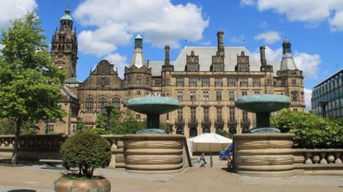 An image of Sheffield Town Hall, a multi-storey sand-coloured building with various small towers on top of the building. A tall clock tower is on the left. Two fountains flank a staircase leading down to the square in front of the hall.