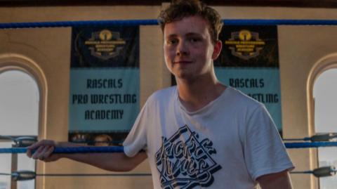 A young man in a white t-shirt standing in a boxing ring smiling.