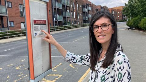 A woman, Nursen Smith, wearing a white top with black patterns on it. She is pointing at a bus timetable on a post.