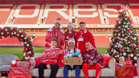 Forest players from the men's and women's first team are sat on a sofa with head coach Nuno Espirito Santo. They are wearing red festive Forest attire and Nuno is holding a basket of donations. There is a Christmas tree to the right and a large wreath to the left. The photograph is inside Forest's City Ground stadium.