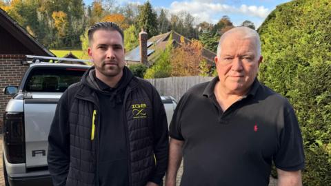 Steve Piller, with black hair and a beard and dressed in black, stands in front of his truck and next to his dad, Simon, who has short white hair and is wearing a black t-shirt.