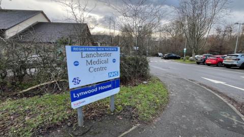 A sign reading Lanchester Medical Centre on the side of a pavement leading to a car park.