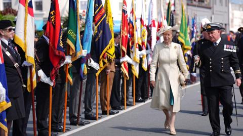 Princess Anne walks past a flag parade as part of Guernsey's Liberation Day celebrations. she is wearing a cream dress. A member of the military stands next to her. 