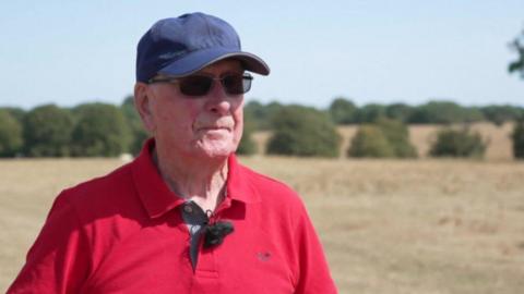 A man wearing black sunglasses and a navy cap is wearing a red polo shirt and looking to the side with a serious expression. The background is blurred but you can make out fields of dry pasture and some trees.