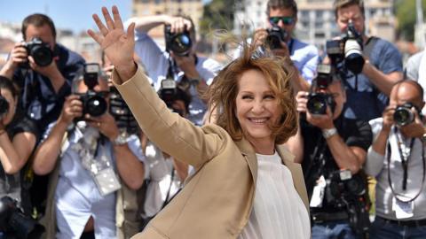 Nathalie Baye, a woman with short brown hair, waves in front of a crowd of photographers in Cannes. She wears a camel-coloured blazer and a white t-shirt. She is smiling.