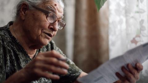 Older woman wearing glasses looks at a bill with curtains and net curtains behind her.