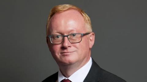 A close-up picture of Luke Akehurst. He has short red hair and is wearing dark-rimmed glasses. The Houses of Parliament can be seen out of focus behind him.