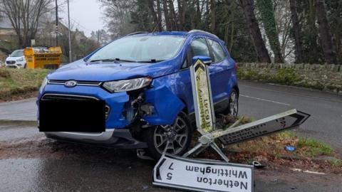 A blue car that has been smashed at the front with the front wheel at an angle at a junction. Beside it is a road sign which has been knocked over. There is road behind it with a stone wall at the side. There is a grass verge to the left and a white van and yellow skip parked behind it.