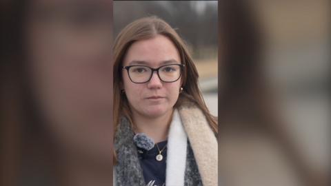 A young woman with glasses and long brown hair looks at the camera