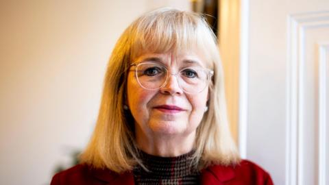 Lady Elish Angiolini KC is stood in a light room and has shoulder length blonde hair. She is wearing a red jacket and a tartan shirt underneath it. She is wearing glasses with transparent frames.