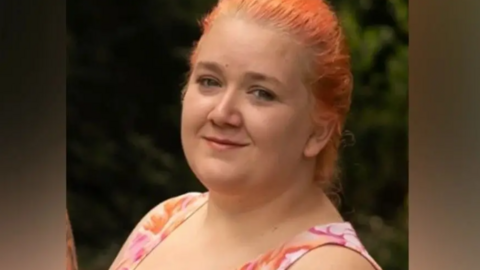 A woman with light red coloured hair looks towards the camera, smiling. She is wearing a top with a floral type design on the shoulder straps