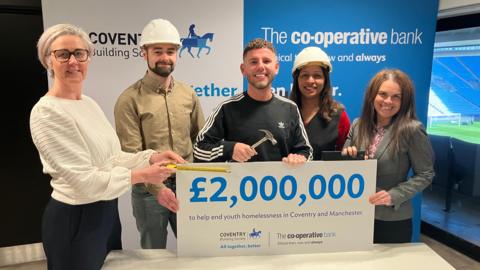 Three men and two women in a room wearing hard hats and holding a tape measure and a hammer stand smiling in front of a large white board which says "£2,000,000 to help end youth homelessness in Coventry and Manchester."