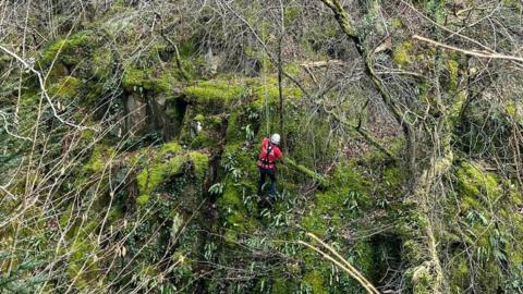A cave rescue volunteer, wearing a red waterproof jacket, black waterproof trousers, black safety harness and a white hard hat abseils down a green mossy rock face.