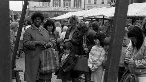 People stand at a busy outdoor market, with stalls, shoppers, and buildings in the background.
