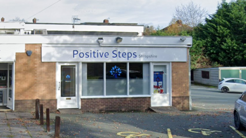 A small building with a white shop front and large glass windows. There is a blue tree decal on the windows, and the sign reads "Positive Steps Shropshire" in blue writing. There are two disbaled parking bays outside it, and a car park around the side