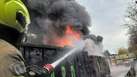 A close up of a fire fighter directing a jet of water on to two shipping containers, with flames and smoke billowing up behind