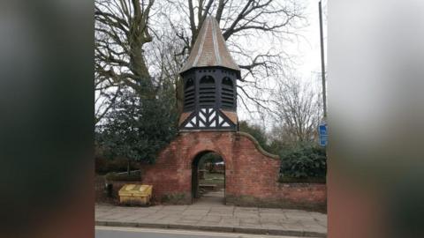 A brick and timber gateway with arched entrance and timber black and white panelling detail and a pointed roof, with trees and shrubs around it and a pavement and part of the road in front just visible